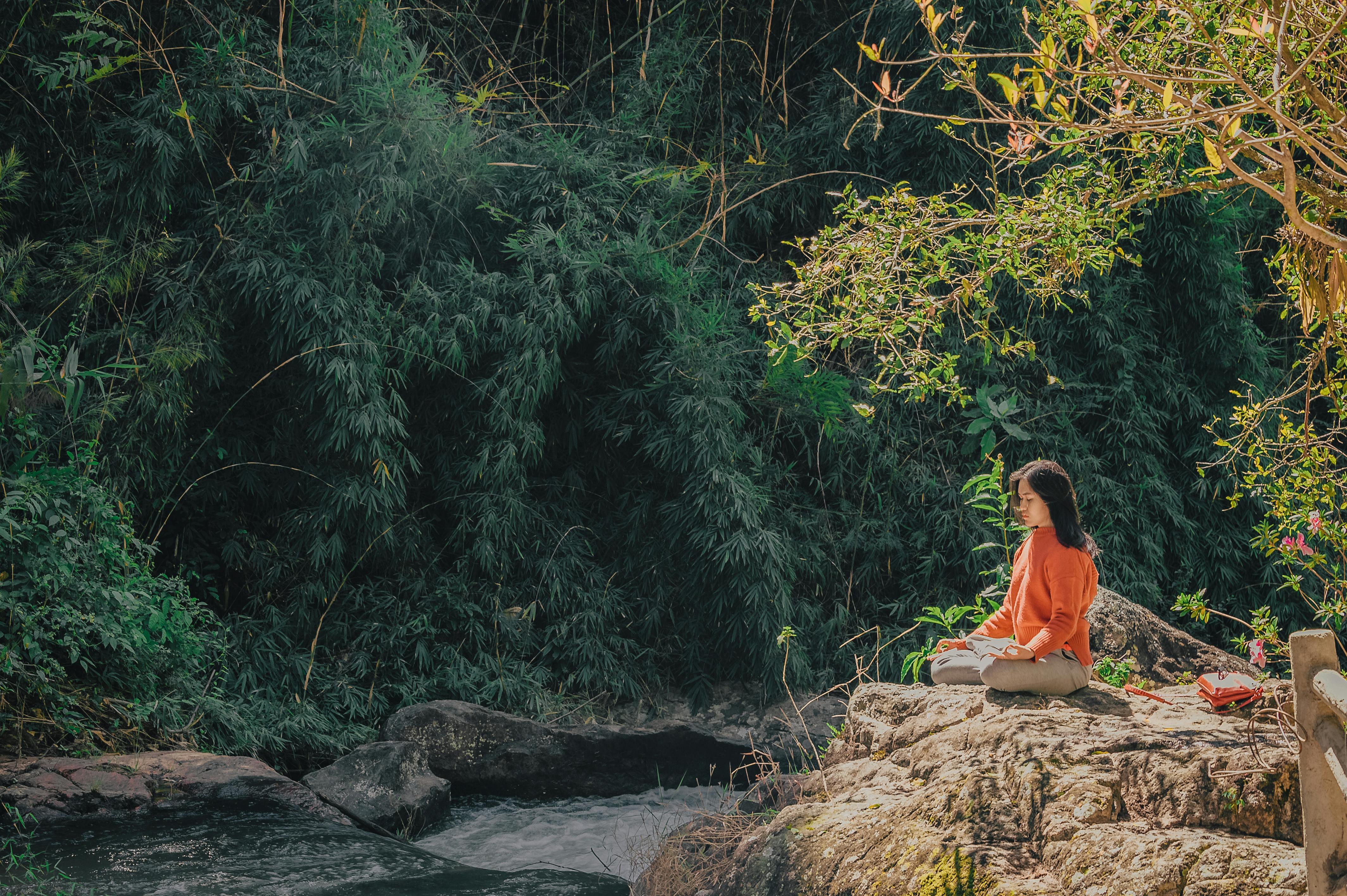 Girl in orange shirt meditating besides a rivulet with bamboo forest in the background